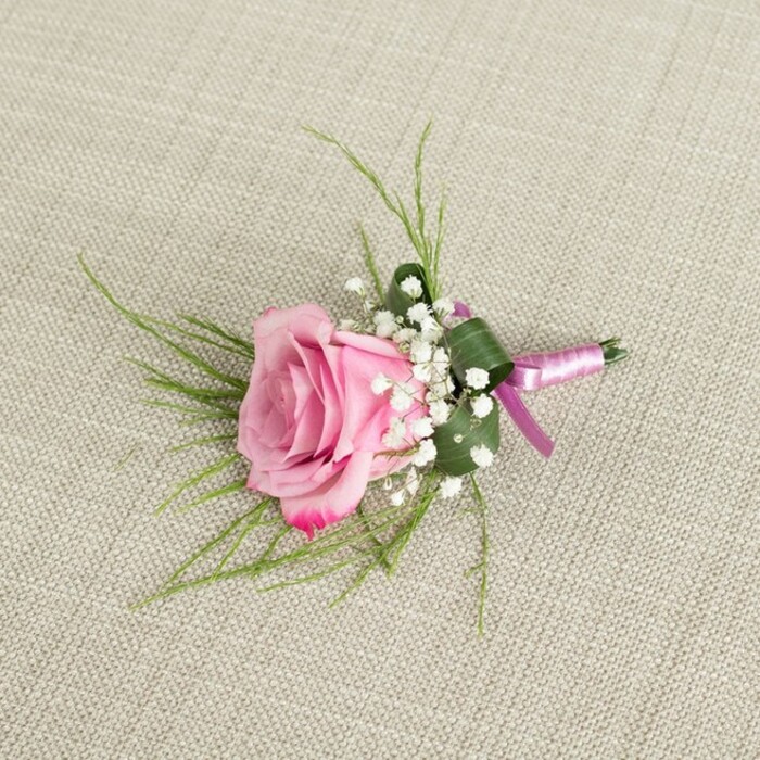 On a softly textured, light beige linen surface that evokes the relaxed elegance of a country-style venue near Epping Forest, a beautifully crafted pink rose boutonniere is laid out and photographed from slightly above. The central rose is in a semi-open state, its petals layered in a graceful spiral that begins with a gentle blush at the core and slowly deepens into a more vivid pink along the edges. The outer petals curl delicately, suggesting a flower cut at peak freshness, with a velvety finish that seems almost touchable. Encircling the base of the rose, clusters of airy white baby's breath form a delicate collar, each tiny bloom adding a soft, cloud-like texture that contrasts with the smoothness of the rose and breaks up the stronger colour. Behind the flowers, slender, grass-like strands of vivid green foliage fan out in an informal, slightly windswept pattern, introducing lightness and a sense of natural movement, as if picked from a garden near Bell Common. A broader, deeper green leaf is carefully folded and wrapped snugly around the stems, lending structure and a tailored appearance, much like the sharp lapels it is designed to adorn. The stems are bound with a lustrous satin ribbon that blends lavender-pink with richer magenta-purple tones, the two shades subtly twisting together and tied into a simple, secure knot. The ribbon's sheen catches the gentle, even lighting, which also casts soft shadows that give depth to the petals and the linen's woven texture. Crafted in the thoughtful, polished style favoured by couples marrying around Epping, this boutonniere feels perfect for a groom, groomsman, or prom guest seeking a romantic yet timeless floral accent.