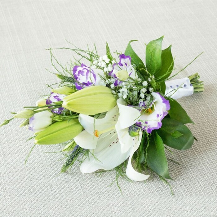 A refined hand-tied bouquet is positioned diagonally across a light beige, subtly textured fabric, the gentle weave of the material evoking the look of natural linen or soft burlap laid out on a rustic table. The eye is immediately drawn to the large white lilies that dominate the centre of the arrangement; their petals are wide and smooth, curling delicately at the edges, with faint freckles and a cluster of golden-yellow stamens at the heart of each bloom. Their presence suggests a rich, sweet fragrance, the sort of scent that might linger in an Epping living room after a same-day delivery. Interspersed among these lilies are several vibrant green buds, still tightly closed and elongated, providing a fresh, youthful energy and promising that the bouquet will continue to evolve over the coming days. Around and between the lilies, Eustoma (Lisianthus) flowers add a touch of drama and sophistication: their petals are ruffled and slightly flared, predominantly white but strongly edged with hues ranging from soft lavender to deep royal purple. Fine, airy clusters of white baby's breath are tucked in between the larger flowers, softening the transitions and giving the arrangement a dreamy, cloud-like texture. The greenery is varied and lush-broad, glossy dark green leaves create a structured backdrop, while finer, more delicate fronds and a few spikier greens push outward, breaking the outline and adding dynamic movement. At the narrow right-hand end, the gathered stems are wrapped in a neat band of white ribbon, subtly embellished with tiny bead-like accents that catch the light, hinting at a bridal or special-occasion purpose. The gentle, diffused lighting and neutral backdrop allow every shade of white, purple, and green to stand out clearly, making this bouquet ideal for wedding photography, engagement celebrations, or elegant home styling in Epping and the surrounding villages.