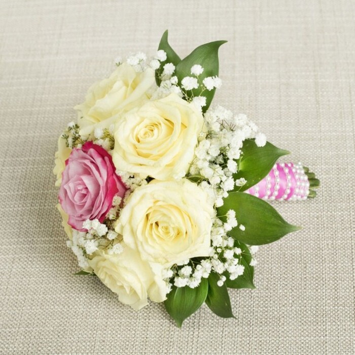 Viewed from above, this compact bouquet, likely destined for a bridesmaid or a spring bride in Epping, rests serenely atop a neutral, linen-textured canvas-an inviting backdrop that highlights every detail. The composition is dominated by a circle of creamy white and pale yellow roses, their petals gently unfurling to reveal a youthful freshness. Offset slightly to the left, one vivid fuchsia-pink rose draws the eye-a dramatic focal point amid the softer blooms. Sprays of delicate white gypsophila are liberally intertwined, imparting lightness and offering a contrast to the velvety fullness of the roses. Framing these blossoms, broad and glossy green leaves evoke the lush edges of Epping's nature reserves and town gardens. The bouquet is carefully finished with a bright fuchsia-pink satin ribbon, its tailored wrapping interrupted by an elegant spiral of small, iridescent pearls. These details combine for a refined and classic look, perfect for Epping wedding celebrations or elegant events at local venues. The bouquet's subtle perfume, mingling rose and fresh foliage, evokes images of spring mornings by Epping Forest. Its artful presentation and intricate details celebrate both modern flair and local tradition-ready to add a touch of romance and joy to any special Epping occasion.