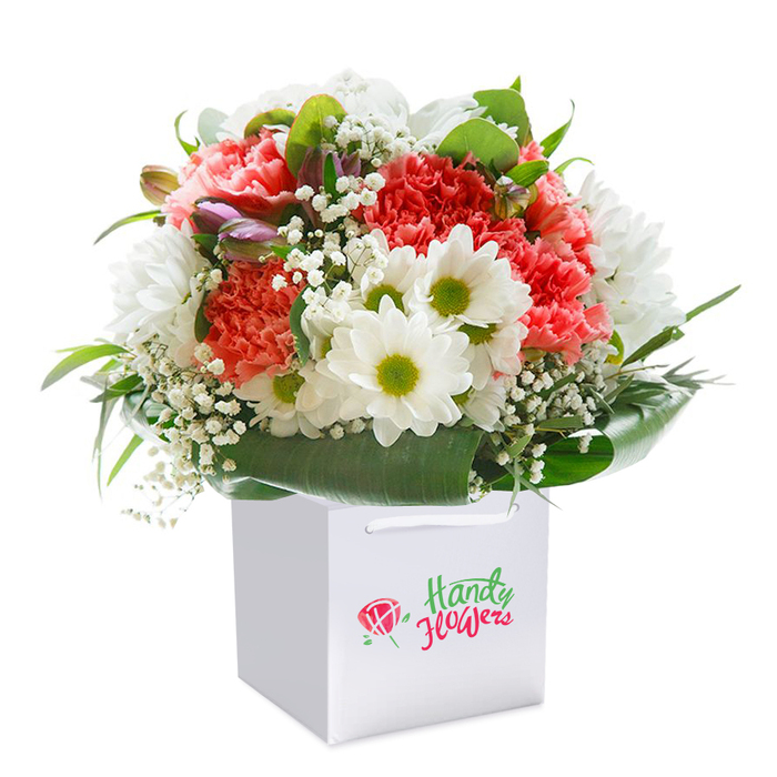 A celebration of the natural beauty found in and around Epping, this professionally arranged floral bouquet exudes elegance, care, and a vibrant sense of occasion. The design is built around a harmonious blend of coral, peach, and red carnations, their plush, ruffled petals forming lush mounds of texture and colour. Interspersed with these bold blooms are crisp white daisies crowned by bright yellow-green eyes, offering a joyful contrast that captures the spirit of community gatherings on Epping's historic streets. Tiny, cloudlike white sprays of baby's breath lend an ethereal quality, their delicate blossoms acting as both filler and focal highlight. On the left, slender stems with purple-tinged buds-perhaps the promise of alstroemeria or another seasonal favourite-peek gently, introducing interest and nuance. Layered beneath, a generous wrap of deep, glossy green foliage encircles the ensemble, echoing the leafy canopies of Epping Forest and providing a structured base. All florals are nestled in a pristine square white gift bag with sturdy ribbon handles, its minimalism underscoring the bouquet's freshness and modern appeal. Against a pure white backdrop, every texture and hue is magnified, turning this bouquet into a timeless choice for birthdays, anniversaries, or as a heartfelt gesture delivered anywhere in Epping. It's a portrait of thoughtful floristry, reflecting local craftsmanship and a keen sense of delight.