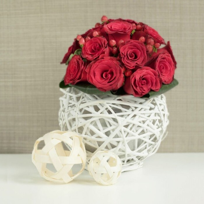 The image presents a carefully styled floral centerpiece that exudes classic romance with a modern twist, the sort of arrangement you might find on a wedding table set in one of Epping's country venues or a chic restaurant near the High Street. At its heart is a closely packed dome of rich red roses, each bloom with petals that appear soft and velvety, ranging from partially unfurled buds to fully open blossoms revealing layered spirals. The roses are arranged so densely that they create a seamless cushion of crimson, catching and holding the light. Threaded among them are clusters of small, glossy Hypericum berries, also deep red but with a slightly different tone and sheen, introducing a glimmering, bead-like texture between the larger petals. Supporting the flowers is a low base of dark green foliage, just visible where the blooms meet the container, its cool tones quietly balancing the warmth of the reds. The arrangement is set in a striking spherical vase made from slim, white-painted twigs or wicker, woven together into an open, nest-like lattice. Through the gaps in this white sphere, shadows fall delicately onto the smooth, bright white tabletop beneath, adding depth and a sense of lightness. To the left of the main piece, two decorative natural-wood spheres rest on the same surface, their pale, beige woven strips echoing the design of the vase but in a softer, untreated tone. The background is a softly blurred wall in a neutral beige or taupe, with a subtle suggestion of fabric or textured wallpaper. The overall atmosphere is calm and sophisticated, yet full of warmth, perfectly suited for a romantic meal at home in Epping or a refined celebration where flowers are meant to feel both contemporary and deeply personal.