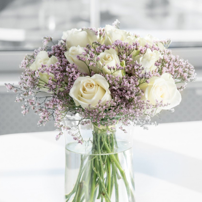 A refined and tranquil floral arrangement fills this photograph, evoking the hush of a thoughtful condolence visit in Epping. At the centre of the shot, a cluster of creamy white roses rises in soft, rounded shapes, each flower fully open and revealing the elegant swirl of its inner petals. Their colour is not a stark white but rather a warm, ivory-tinged cream, lending the bouquet a gentle, comforting warmth instead of formality. Surrounding and weaving through these roses are plentiful sprays of tiny lavender-pink filler flowers, likely statice, whose many small florets give the impression of fine lace or delicate mist. This haze of pastel purple tones adds texture and depth, softening the outlines of the roses and creating an almost cloud-like effect that feels soothing to look at. The entire bouquet sits in a clear, cylindrical glass vase, simple in design so as not to distract from the blooms. Inside, the long green stems are neatly cut and arranged, visible through the transparent glass and disappearing into fresh, clean water that catches small glimmers of reflected light. The vase is placed on a smooth white surface, perhaps like a polished table in a quiet Epping home, which reflects a touch of the arrangement's light and colour. In the background, everything is softened into pale, neutral tones, with no distinct details, allowing the arrangement to stand alone as the clear focal point. The lighting is gentle and diffuse, reminiscent of natural daylight filtering through a nearby window off Epping High Street, and it enhances the bouquet's role as a symbol of remembrance, condolence, and quiet reassurance during times of loss.