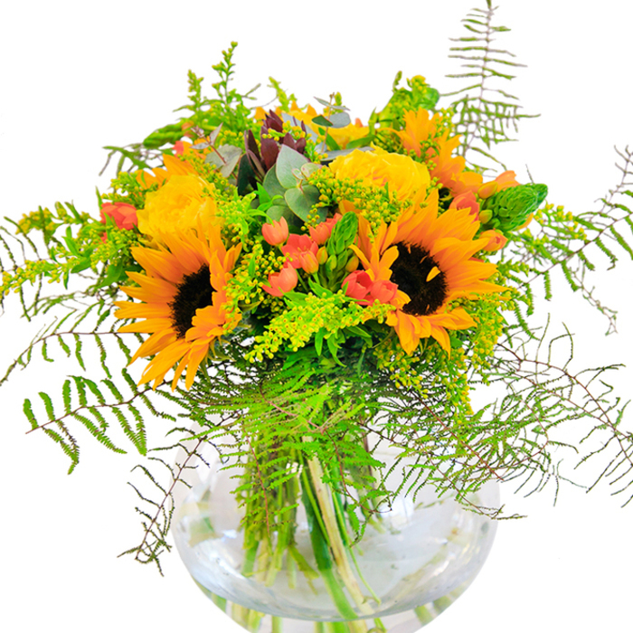 Upon first glance, this abundant floral arrangement seems to capture all the brightness of an Epping summer's day, with sunflowers and yellow garden roses beaming together in perfect harmony. The generous, ruffled petals of the sunflowers evoke a sense of golden sunlight streaming across local fields, while classic garden roses unfurl in tightly layered spirals, their soft yellow tones and subtle fragrance adding layers of elegance. Scattered between, vivid red Hypericum berries punctuate the arrangement with bursts of vivacity, like joyful laughter echoing across the village green. Fern fronds tumble gracefully from the edges, their feathery softness mimicking the gentle movement of wind along York Road, contrasting against bold flower forms. Wispy sprays of goldenrod and slender greens bring delicate texture, while eucalyptus leaves in muted blue-green hues lend a refreshing note suggestive of peaceful Epping woodland walks. Everything comes together in a clear, round glass vase, where fresh stems submerge in crystal water, amplifying the arrangement's lively, welcoming feel. The thoughtfully balanced colors and natural textures make this bouquet an ideal choice for celebratory moments, housewarmings in Coopersale, or simply bringing the spirit of the outdoors in. Each bloom and berry is arranged with artful care, transforming a simple space into a celebration of Epping's vibrant floral and community life.