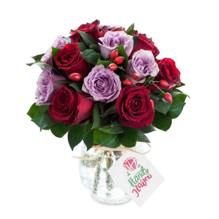 A carefully composed bouquet of rich burgundy and soft lilac roses sits elegantly in a vintage-effect mercury glass vase, the colours intensified against a bright white background. The arrangement is compact and rounded, with deep red and crimson roses clustered closely alongside pale purple and dusty lavender roses, each bloom open to reveal velvety layered petals and delicate unfurling textures. Interspersed among the roses are dense sprays of dark green foliage-likely Ruscus or Salal-whose cool, glossy leaves provide depth and a natural frame for the flowers. Bright red-orange rose hips or hypericum-like berries are artfully placed throughout the bouquet, offering a lively contrast in colour and a tactile, rustic element that complements the roses' elegance. The vase, with its bulbous base and slightly tapered neck, shows a silvered, crackled mercury finish that shimmers gently in reflected light; a simple strand of natural twine or raffia is tied around the neck, suspending a small rectangular white tag decorated with a tiny stylised rose sketch and a handwritten-style sentiment in muted green and pink. As prepared by a local shop familiar with Epping's tastes-Flowers Epping-this bouquet conveys romance and thoughtful generosity, ideal for delivery along the High Street, to homes near the tube station, or to country-edge cottages by Epping Forest. The soft studio lighting highlights the contrasts of texture, scent, and colour, suggesting a fresh, premium gift for anniversaries or heartfelt celebrations.