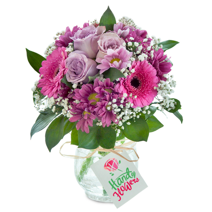Centred against a clean, white background evocative of a modern studio, this lovingly arranged bouquet reflects the natural beauty and abundant spirit of Epping. It bursts with a harmonious palette of soft lavender, fuchsia pink, and deep plum, illuminated by cool accents of crisp white and verdant green. Elegant pale lavender roses form the tranquil heart of the bouquet, their petals layered in intricate spirals that seem almost sculpted, suggesting quiet grace. Bold gerbera daisies in electrifying pink, their wide petals drawing the gaze, stand at attention nearby, each with a velvety dark centre. Clusters of magenta and plum chrysanthemums create depth and a sense of wild textural variety, akin to blooms gathered fresh after a walk through the Epping Forest. Generous tufts of baby's breath gently hover between the larger blossoms, their tiny white flowers adding a dreamy, airy lightness. Draped throughout, rich green foliage frames and supports every bloom. The arrangement sits elegantly in a distinctive clear glass vase with a unique cracked-ice pattern, the changing light casting interesting reflections across the surface. Tied around the vase is a simple rustic raffia ribbon, accented by a small rectangular gift tag adorned with a delicate pink rose illustration. This bouquet is perfectly suited for express delivery across Epping-whether as birthday greetings, an anniversary surprise, or an everyday gesture of gratitude-and its fresh, vivid presence recalls the local countryside, welcoming any recipient with a sense of seasonal abundance and skilled craftsmanship.