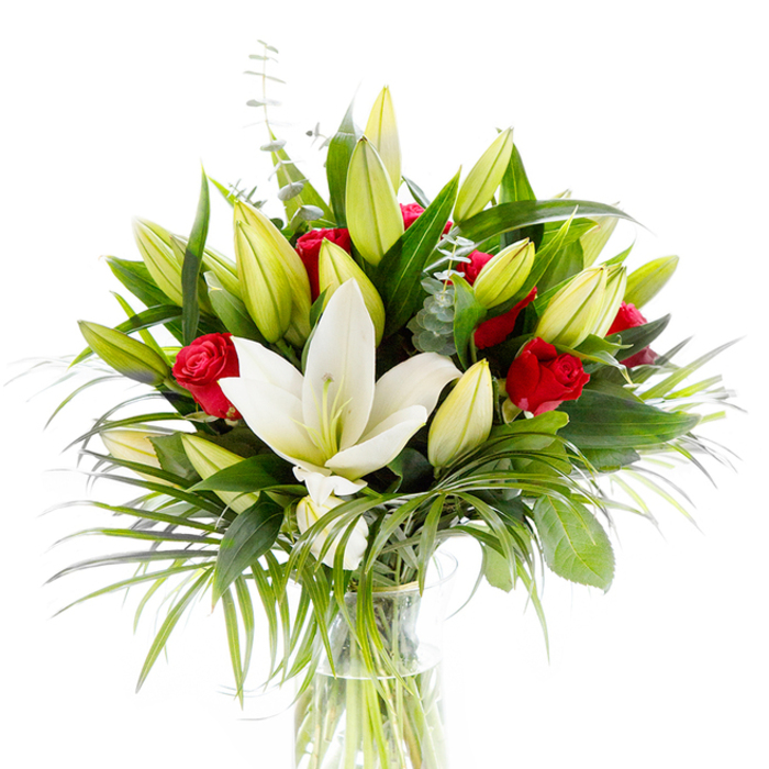 A striking, gallery-like floral arrangement presented in a clear glass vase, photographed against a bright white background that isolates its layered textures and vivid colours. Central to the display is a single, fully open creamy Oriental lily whose smooth, slightly curved petals form a luminous, almost sculptural bloom; at its heart delicate pale yellow stamens and a light green pistil create a quiet point of focus. Surrounding this are many lily buds in soft lime-green and creamy-yellow tones, some tightly closed and others beginning to unfurl, suggesting gentle motion and future blossoms. Interwoven among the lilies are several small red roses and rosebuds with velvety, deep crimson petals still curled inward, their passionate hues providing bold contrast and classic romance. The bouquet is richly populated with varied greenery: broad, dark green lily leaves frame the composition and give it solidity, while long, feathery palm fronds spill outward in lighter emerald tones that add rhythm and a subtle tropical note. Sprigs of silvery eucalyptus with their characteristic rounded leaves are threaded through the arrangement, introducing cool, muted highlights and a soft texture against the glossier foliage. Through the transparent vase, tidy green stems sit in clear water, a simple detail that emphasises freshness and professional care. The controlled studio lighting enhances the lilies' creamy sheen and the roses' velvety depth, conveying a mood of refined celebration. This elegant blend of white lilies and red roses reads as a perfect gift for anniversaries, birthdays, or occasions calling for a sincere expression of admiration and love.