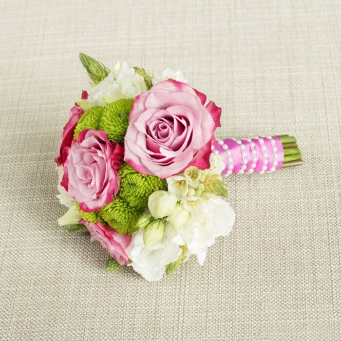 A neatly composed, small bouquet is shown resting horizontally on a light beige fabric with a visible weave, reminiscent of natural linen laid out on a table in a home near Epping Forest. The neutral backdrop acts like a calm canvas, allowing the colours and textures of the flowers to come forward. At the centre, two generous pink roses dominate the scene, their petals opening fully to reveal intricate, layered spirals. The rose colour begins as a soft blush in the middle and gradually deepens to a richer fuchsia along the outermost petals, where hints of magenta at the tips create a sophisticated ombré effect. Clustered closely around these roses are bright lime-green button chrysanthemums, each head a tight, rounded cushion of minuscule, densely packed petals. Their vivid green tone injects energy and youthfulness into the arrangement while also enhancing the freshness of the pink roses. Threaded between the larger blooms are airy white and creamy filler flowers-likely small lisianthus, freesias, or waxflowers-with starry blossoms and occasional tiny yellow eyes that add delicate intricacy. Subtle touches of deep green foliage peek from underneath, creating a gentle frame and natural depth. The stems of the bouquet are cut short and bound together in a compact handle, wrapped smoothly in fuchsia satin ribbon that gleams softly under the even light. Over the ribbon, a fine strand of white faux pearls is wound diagonally, as though a piece of jewellery has been gently draped around the handle, giving a classic bridal refinement. The entire composition suggests a bridesmaid's posy or petite bridal bouquet, ideal for ceremonies at local churches in Epping or intimate celebrations at country venues along the edges of the forest, evoking romance, joy, and thoughtful craftsmanship.