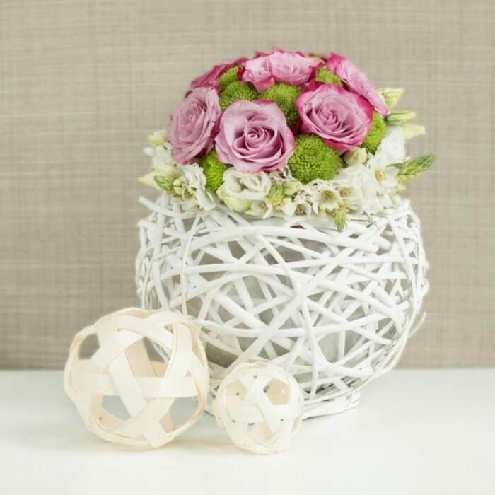 This image presents an elegant, wedding-ready table arrangement styled in a minimalist studio setting, ideal for a refined celebration in Epping or a romantic dinner at home. The focal point is a round, white woven vase, crafted from slim rattan-style twigs interlaced into a spherical, open design that feels light and sculptural. Slightly flattened at the base, the vase rests securely on a pristine white surface, its pale tone echoing the clean simplicity of the tabletop. Rising from this vessel is a lush, carefully domed bouquet made up mainly of roses in soft pink and gentle lavender hues, some petals edged with a richer pink that adds depth and visual interest to each bloom. The roses are intermingled with bright green button chrysanthemums whose small, tight clusters create a lively pop of colour and a firm, tactile texture. Nestled low around the base, dainty white filler flowers, possibly baby's breath or tiny spray roses, frame the central blooms with a whispery softness that makes the overall shape feel full yet airy. To the left of the vase sit two decorative orbs made from pale natural wicker or wood, one larger than the other, each formed from thin strands woven into open, geometric patterns. Their natural beige tones gently contrast with the white vase and tabletop while still harmonising with the neutral, linen-like beige background. The lighting is gentle and diffuse, similar to early afternoon light filtering through windows on Epping High Street, illuminating the arrangement without harsh shadows and highlighting every curve of petal, every twist of woven twig, and every carefully considered detail crafted by an experienced local florist.