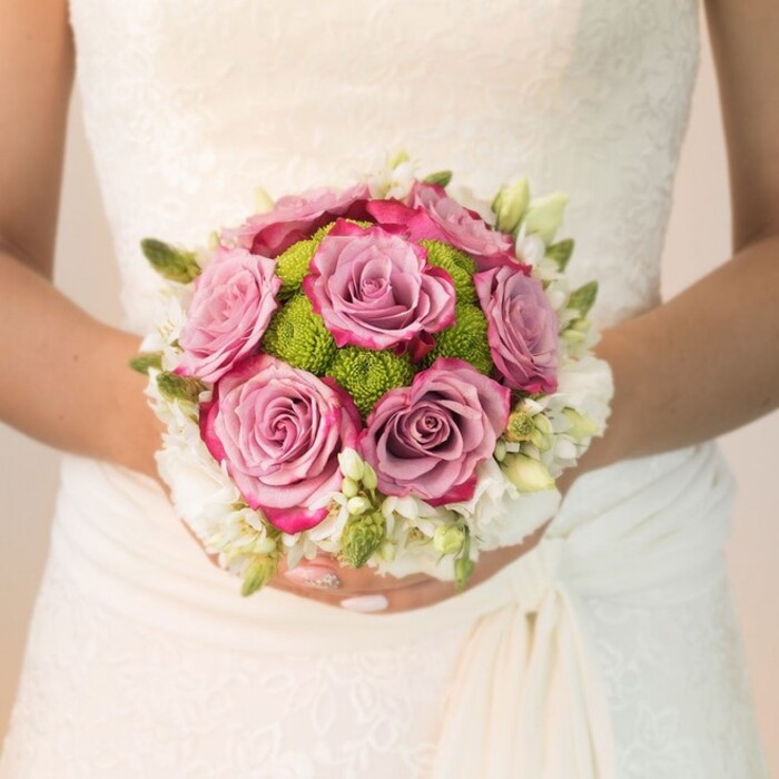This evocative bridal portrait captures a waist-level glimpse of a bride in Epping as sunlight softly bathes her lace-covered ivory gown, lending a glow to every delicate detail. Gracefully poised, her manicured hands-nails painted the palest blush, with a jewelled accent on one finger-support a perfectly round, exquisitely designed bouquet. The arrangement is a study in romance and texture: at its heart lie several open mauve roses, their plush petals shifting from smoky lavender to vivid fuchsia along the edges, creating a painterly display. Cupped around these are tight orbs of lime-green button chrysanthemums, their vibrant hue and textured surface adding local charm and freshness, recalling the lively energy of the Epping market. Encircling this core are wisps of white blooms, likely freesia or petite spray roses, complemented by fresh green buds and subtle foliage, infusing the bouquet with lightness and understated elegance. The bride's dress forms a subtle, intricate backdrop-the lace bodice and softly gathered skirt reflecting Epping's blend of tradition and contemporary style. The overall composition radiates a calm, serene joy, making this bouquet a showpiece of local wedding floral craftsmanship. Its harmonious mix of pastels, vibrant greens, and luminous whites captures the spirit of a joyful celebration in the heart of Epping.
