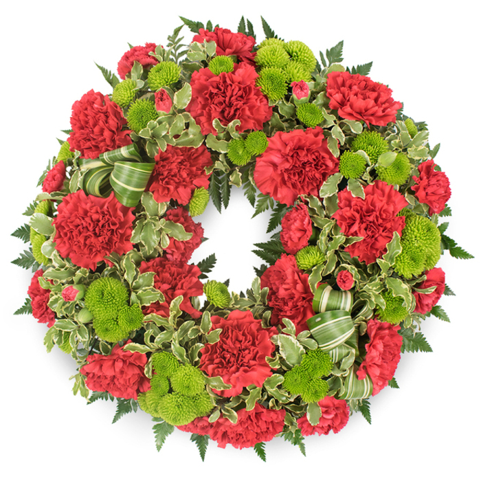 A traditional circular funeral wreath, densely packed with deep red carnations and vivid lime green button chrysanthemums, is displayed against a bright white background, highlighting every ruffle, leaf, and petal as though lit by soft daylight through an Epping chapel window. The ring is completely covered in flowers, leaving no gaps, giving it a substantial, comforting presence. Red carnations form the main body of the arrangement, their multi-layered petals folding and crinkling into rich, velvety rosettes that flow seamlessly around the circle. Some carnations are in full bloom, with wide open faces, while others are partly unfurled, adding depth and nuance to the design. Between these crimson clusters, neat, spherical pompon mums in a striking lime green pop forward, their tidy, button-like heads made of tightly packed, tiny florets. This fresh green is bright but not harsh, offering a sense of gentle renewal beside the more solemn red. Encircling and weaving through the flowers is a lush mantle of mixed foliage. Dark green ferns add a light, feathery texture, while variegated pittosporum leaves, with their cream-edged margins, bring soft highlights and a slightly mottled pattern that keeps the eye moving around the wreath. A coordinating ribbon, striped in different shades of green, is nestled in folded loops among the flowers, its fabric curves echoing the circle and suggesting the careful hand of an experienced florist. The overall impression is of a respectful and vibrant tribute, suitable for funeral and remembrance services throughout Epping, from intimate family gatherings at home to formal services in local churches and crematoria, where a classic red-and-green wreath can quietly express enduring love and farewell.