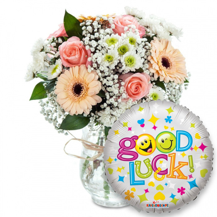 This image shows a warm, encouraging Good Luck set arranged with care, presented against a plain white field so every colour and texture reads clearly. At the upper left a luscious bouquet fills a clear glass vase, the neck finished with a rustic twine knot that hints at artisan hands. The flowers include soft peach Gerbera daisies with deep brown centres that draw the eye, and pale pink roses whose layered petals suggest a gentle perfume. Interlaced throughout are clouds of baby's breath - tiny white points that soften stems like delicate lace - and clusters of white and lime-green chrysanthemums that bring bright, crisp punctuation to the arrangement. Dark green foliage slips between blooms, giving the composition depth and a fresh, garden-picked feel. Dominating the foreground to the right is a shiny silver Mylar balloon, its surface reflecting light and dotted with colourful shapes: blue and pink hearts, yellow and blue stars, and tiny colourful crosses. Across it runs the jubilant message Good Luck! in a bouncy, multi-hued type; playful yellow smiley faces replace the o's in Good - one winking, one making a silly face - making the sentiment feel personal and affectionate. The lighting is soft and even, showing petal texture and the balloon's metallic gleam without harsh shadows. This pairing reads as a reassuring gift for exams, job interviews, farewells or fresh starts, the sort of hand-tied bouquet and balloon a local customer might pick up before a walk through Epping Forest or a visit to the station on the High Street.