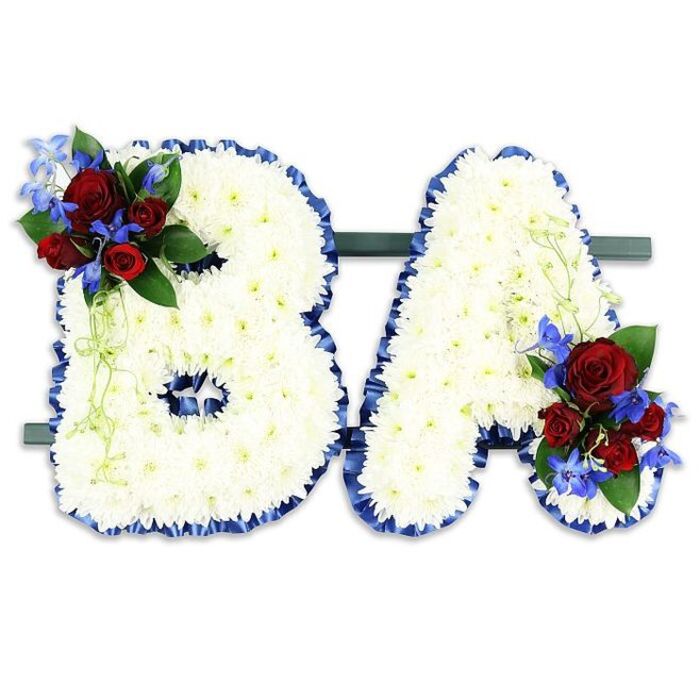 This meticulously arranged memorial pair spells the capital letters B and A in a traditional sympathy style that balances formality with tenderness. Each letter is densely covered in hundreds of small, snowy-white chrysanthemum florets, layered to form a pillowy, cloud-like texture that suggests peace and purity. The silhouette of each character is crisply outlined with a ruffled royal blue satin ribbon, its cool sheen delineating the shapes and adding a formal, respectful frame. Nestled into the upper left curve of the 'B' is a concentrated cluster of deep red roses, offering a velvety warmth, accented by upright, intensely blue delphinium spikes and a mix of dark green foliage and pale trailing tendrils. The 'A' is similarly embellished with a mirrored cluster at its lower right, creating visual harmony and a deliberate, comforting rhythm of colour. Both letters are presented side-by-side on a discreet dark-green rectangular stand that stabilises the display and suits placement in chapels, funeral halls or beside a memorial plinth. The composition sits against a neutral white background that allows the reds, blues and greens to read with clarity. Often chosen for funeral services and commemorations around Epping - from the Market Square to the quieter lanes edging Epping Forest - this piece conveys respect, personal connection and a steady calm through considered floristry and classical symbolism.