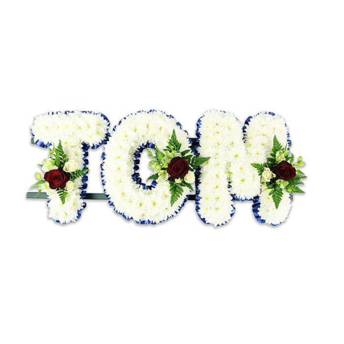 This personalised floral tribute spells TOM in large, three-dimensional lettering entirely covered in small, immaculate white chrysanthemum-like blooms, their rounded heads packed close to form a smooth yet textural surface. Each letter is edged with a bold royal blue ribbon that frames the white flowers with crisp, elegant contrast, bringing a touch of solemn dignity to the composition. Placed thoughtfully within the centre or lower portion of each letter is a compact spray: a single deep red rose takes centre stage in every cluster, its rich colour and soft, velvety petals a clear emblem of love and remembrance. Accompanying the red roses are smaller creamy-white spray roses or buds and slender green fern fronds, whose fine fronds and glossy leaflets add movement and depth against the chrysanthemum backdrop. The letters are subtly mounted on a dark green or black metal framework and connected by a discrete horizontal bar visible at the base, designed to hold the arrangement upright during a funeral or memorial service. Photographed on a clean white background under soft lighting, the textures and colours read sharply-the blue ribbon gleams, the white blooms feel cool and tender, and the red rose centres draw the eye. The overall impression is composed and respectful, suited for display at a chapel, crematorium or family wake in Epping, perhaps brought from a local workshop close to the station and Bell Common. It conveys quiet dignity: handcrafted, balanced and meant to comfort mourners through well-chosen florals and measured, professional design.