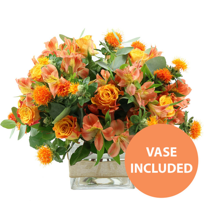 An abundant bouquet in glowing shades of orange, yellow, and peach is captured against a bright white backdrop, every flower sharply defined as if freshly arranged in a local Epping florist's studio. Dominating the center are velvety orange roses, their petals spiralling outward in rich, saturated tones that gently transition toward warm yellow nearer the heart, giving each bloom a sense of living movement. Among them nestle sunny yellow roses and garden-style blossoms, tightly petalled at the core and delicately ruffled at the outer edges, adding a sense of fullness and classic elegance. Woven through these larger flowers, clusters of peach and light orange alstroemeria bring a softer, more delicate note; their slender petals curl slightly at the tips and reveal the characteristic speckled and striped pattern on the inner surfaces, as if they have been lightly painted. Adding a striking textural accent, small rounded flowers with spiky, radiating petals in an intense orange hue are scattered throughout the bouquet, echoing the wild charm of hedgerow thistles along the lanes around Epping. Surrounding and supporting the blooms is a generous collar of greenery, including broad, silvery eucalyptus leaves and deeper green foliage, which adds both structure and a cool contrast that makes the warm colours appear even more vibrant. The bouquet stands in a clear, square glass vase, its clean geometry contrasting with the natural softness of the flowers. Around the central section of the vase is a band of natural beige burlap or jute ribbon, wrapped neatly to introduce a rustic, handcrafted element that would suit a home near Epping Forest or a cosy café off the High Street. In the lower right corner of the image, a vivid orange circular graphic displays the words VASE INCLUDED in bold white capitals, indicating that this arrangement arrives as a complete, ready-to-display floral gift, ideal for birthdays, thank you gestures, or seasonal celebrations across the Epping area.
