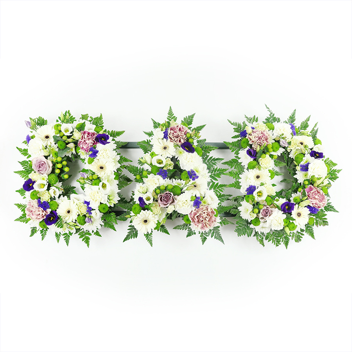 The image presents a touching floral word tribute that clearly spells DAD in three side-by-side capital letters, laid out in a straight horizontal line and photographed against a bright, uncluttered white background. Each letter is solidly built from a mass of pristine white flowers, primarily plump chrysanthemums and gerbera-type blooms, their rounded petals forming a plush, textured surface that feels soft and gentle to imagine touching. Among the whites, you can pick out occasional smaller blossoms and buds, bringing subtle variation in shape while keeping the overall look calm and unified. Around the perimeter of each letter, there is a rich edging of vivid green foliage: delicate fern fronds and leafy sprigs that follow every curve and corner, giving the tribute neat definition and a natural, garden-like character you might associate with the green spaces around Epping Forest. Dotted through the letters are accents of deep purple and violet, reminiscent of pansies, lisianthus, or spray chrysanthemums, adding depth and a sense of dignified formality. Softer lavender and pale mauve roses, possibly accompanied by frilled carnations, introduce gentle warmth and evoke tender memories of a much-loved father. The lighting is even and clear, helping each flower head stand out distinctly and ensuring that DAD reads clearly from any angle, as it might when placed atop a coffin or stand at a service in Epping. This arrangement captures the essence of a bespoke funeral tribute, the kind of carefully considered design created by an experienced local florist to express love, gratitude, and respect when words may feel insufficient.