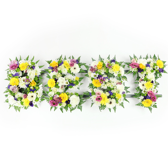 A carefully composed, horizontal floral tribute forms the word BEST in bold capital letters, photographed square-on against an immaculate white background to emphasise colour, form, and texture. Each letter is built from a generous massing of blooms: bright yellow roses and gerbera lend a sunny, buoyant character; pure white, fluffy chrysanthemum-like flowers supply volume and a soft tactile quality; and a range of purples-from lavender lisianthus to deeper violet spray chrysanthemums-introduce an understated, reverent tone. Brimming between these larger focal flowers are lively bursts of magenta-pink spray roses or carnations and delicate pale green and white filler blooms that add intricacy and depth. The perimeter of every letter is edged with dark green fern fronds whose feathery texture frames the composition and contrasts beautifully with the bright petals. In a few shallow gaps the dark green or grey structural bars are visible, a discreet reminder of the skilled engineering beneath the floral surface. The lighting is even and luminous, bringing out the cool whites and warm yellows while preserving the subtle mid-tones of lavender and magenta; the implied fragrance is soft and floral, a calming blend of rose and chrysanthemum. This piece reads as both an affectionate celebration and a solemn tribute-equally at home at a commemorative service near Epping Station, at a neighbourhood memorial in Theydon Bois, or placed on a family mantel as a memory of a cherished life. The arrangement speaks to experienced floristry, thoughtful colour balance, and tactile richness.