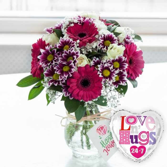 A cheerful, modern bouquet sits proudly on what appears to be a white tabletop, glowing in the kind of bright, diffused daylight that might spill into a sitting room near Epping Forest or along the High Street. The arrangement is a joyful combination of fuchsia gerbera daisies, their rich magenta petals arranged in perfect circles around dark, almost black centres, providing a bold and confident focal point. Surrounding them are lively clusters of chrysanthemums with striking bi-colour petals: deep purple at the core gradually lightening to pure white at the edges, each bloom anchored by a clear yellow centre that adds a burst of sunshine. Interwoven are creamy white roses, some still slightly cupped, others beginning to unfurl, adding a sense of quiet romance and refined elegance. Wisps of white baby's breath drift through the composition like tiny stars, filling spaces softly and giving the bouquet a layered, textured look. Lush green foliage peeks out from beneath and between the blossoms, framing the colours and giving the arrangement a natural, garden-fresh fullness. All of this is gathered into a clear, rounded glass vase, where the green stems stand neatly in clean water. Around the neck of the vase, a piece of rustic, natural-coloured twine is tied in a casual bow, from which hangs a small rectangular white tag displaying a simple pink rose graphic and friendly green script, subtly suggesting that the flowers were hand-selected with care. To the right of the vase, a polished heart-shaped foil balloon with a reflective silver edge and white centre adds a playful finishing touch, decorated with colourful letters that spell Love n' Hugs 24-7. The softly blurred, light-filled backdrop enhances the sense of joy and occasion, making this pairing feel perfect for romantic gestures, birthdays, or surprise deliveries to loved ones around Epping.