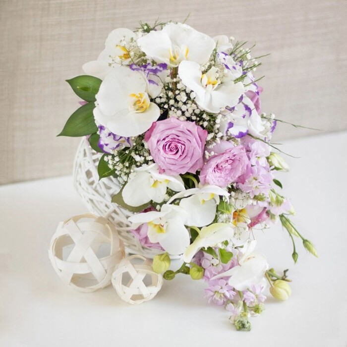 A sophisticated floral arrangement, perfect for a wedding reception or anniversary celebration in Epping, is positioned centre stage on a flawless white surface. The flowers are gathered in an arresting white sphere-shaped vase whose open, criss-crossed weave resembles delicate rattan or artistically wound twine, giving the entire composition a light, airy feeling. Rising from this sculptural base is a generous bouquet dominated by snowy Phalaenopsis orchids, each bloom broad and serene, with creamy petals that curve gently around golden and yellow-flecked centres. These orchid faces tilt outward, lending the design a sense of quiet confidence and modern luxury. Between them, roses in soft blush pink, dusky lavender, and pastel lilac unfurl in plush, many-layered spirals, their velvety texture implying a subtle, powdery fragrance. Smaller accent flowers with white petals edged in shades of purple, reminiscent of Eustoma or Lisianthus, nestle among the roses, adding tonal depth and an extra ruffle of romance. Fine sprays of white gypsophila thread throughout, like tiny stars against a twilight sky, filling the arrangement with an ethereal softness. Light purple stock or similar elongated blossoms flow over the vessel's rim in a gentle cascade, their stems drawing the eye downward and adding movement, while glossy leaves in varied greens frame the pastels with a natural vibrancy. To one side of the main display, two cream-coloured, woven decorative balls-one larger, one smaller-rest casually on the table, their open latticework echoing the vase and tying the scene together. The background, a subtly textured beige wall, remains softly out of focus, allowing every petal and woven strand to stand out, as if prepared by an experienced local florist for a stylish event near Epping High Street or a romantic dinner at a nearby country inn.