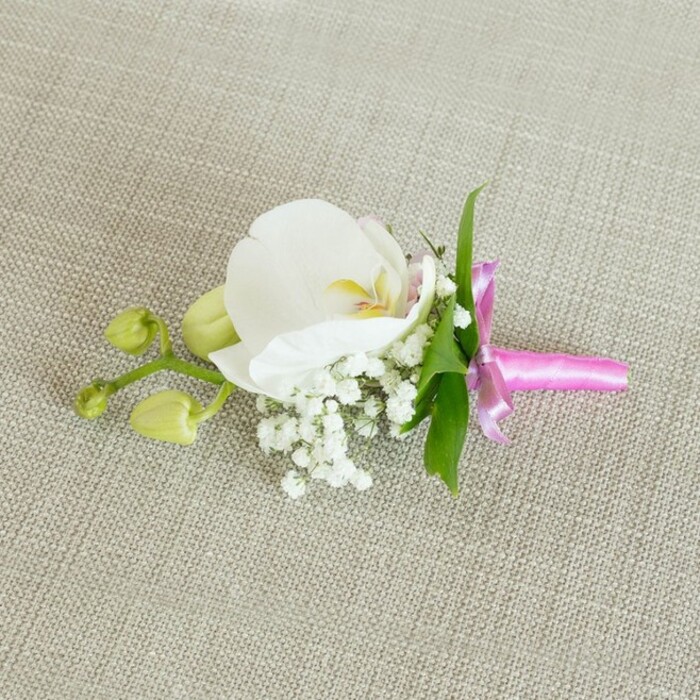 An elegant orchid boutonniere or corsage is laid horizontally on a softly textured, light oatmeal-coloured linen background, the woven fabric forming a calm, neutral stage that enhances the flowers' gentle colours. At the centre of the piece sits a single, flawless white Phalaenopsis orchid bloom, its broad petals gracefully arched and luminous, as though just opened in the cool morning air. The heart of the orchid reveals intricate notes of warm creamy yellow and a faint, blush-pink hue, delicately patterned within the throat of the flower, giving it an almost porcelain refinement. Gathered closely at the base of this focal bloom, small clusters of white baby's breath create a dreamy, cloud-like surround, their tiny spheres and branching stems adding lightness and soft volume. A handful of slim, bright green leaves emerge subtly behind the petals, bringing a touch of garden freshness that feels at home in the leafy streets leading toward Epping Forest. To the side, a short, branching stem showcases a few tightly closed orchid buds in pale lime green, their rounded, unopened forms suggesting new beginnings and the quiet anticipation of future blossoms. The stems beneath are neatly and firmly wrapped with a narrow fuchsia pink satin ribbon, tied into a refined little bow with short, polished tails, its glossy texture catching the light and injecting a joyful accent of colour. The gentle, even lighting highlights every nuance-the velvety orchid, the airy baby's breath, the smooth ribbon, and the subtle grain of the linen. Overall, the image evokes a sense of timeless, romantic elegance, perfect for a groom's lapel, a bridesmaid's corsage, or a formal guest at a local celebration or wedding reception in Epping.