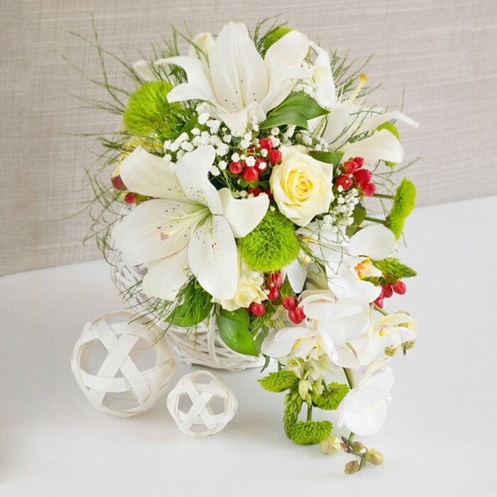 A beautifully composed wedding-style table arrangement is showcased on a bright white surface, backed by a softly textured beige wall that gives the scene a warm, neutral glow. The flowers are gathered low and full in a round white woven basket, its wicker pattern adding a natural, hand-crafted feel that would sit comfortably in an Epping home or boutique venue. Front and centre, several large white lilies unfurl their trumpet-shaped petals, some showing delicate dark speckles and prominent stamens dusted with golden pollen, creating a sense of drama and fragrance. Just off the top centre, a single creamy yellow rose, perfectly formed and slightly cupped, acts as a gentle focal point, its soft colour easing the transition between the pure whites and vivid greens. From the right-hand side of the basket, a trail of white phalaenopsis orchids cascades gracefully downward, each flower with broad, smooth petals and a hint of ivory at the centre, forming an elegant waterfall reminiscent of a bridal bouquet being laid carefully on a registrar's table in Epping. Around these key blooms, the florist has woven in an array of greenery: fine, feathery leaves, structured stems, and bright green pom-pom chrysanthemums that contribute a playful, textural contrast. Tiny white stars of baby's breath thread through the arrangement, while glossy ruby-red berries are dotted here and there, giving small flashes of colour like berries spotted along a woodland path near Epping Forest. To the left of the main basket, two white woven spheres of different sizes sit on the table, echoing the natural textures and reinforcing the airy, contemporary aesthetic. The entire composition radiates polished elegance, freshness, and quiet celebration, perfect for wedding receptions, vow renewals, or intimate dinner parties across Epping.