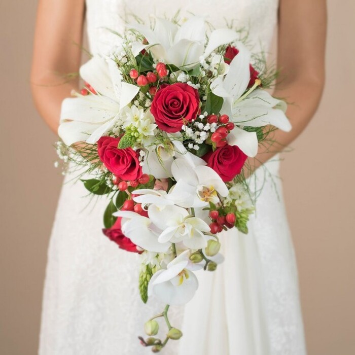 A romantic bridal image conjures the essence of an Epping wedding, where a bride in a classic lace gown holds an extravagant teardrop bouquet crafted by local florists. Framed against a softly blurred beige backdrop, reminiscent of the calming landscapes near Epping Cricket Ground, the composition draws immediate attention to the intricate details of the bouquet. Several pristine white oriental lilies, their broad petals and golden stamens on full display, command central focus, symbolizing purity and classic beauty. Interwoven throughout are lush, deep red roses-their tight, velvety blooms radiate warmth and devotion, echoing the spirit of romantic celebrations along Epping High Street. Tiny white baby's breath clusters and small, bright red hypericum berries pepper the bouquet, infusing it with gentle, playful texture and color. The cascading shape is achieved with the help of long, trailing Phalaenopsis orchids, whose soft, white blossoms and subtle yellow centers add graceful movement, evocative of Epping Forest's winding paths. The bouquet's base of wispy foliage, including airy asparagus ferns and sturdy green leaves, offers a supportive framework for the spectacle of blooms. Held gently in the bride's lightly tanned arms, the bouquet offers a vision of elegance, designed not only to complement her lace-adorned dress but to celebrate Epping's blend of tradition and natural charm. Flowers Epping's artistry truly shines, presenting a centerpiece that speaks of sophisticated love, local heritage, and the enduring joy of wedding day dreams.