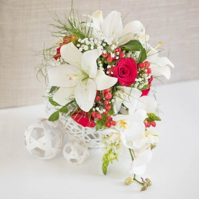 A sophisticated wedding table arrangement is carefully staged against a softly textured beige backdrop, evoking the calm, neutral tones of a boutique venue tucked just off Epping High Street. At its heart is a round, white container crafted in an open, basket-like weave, the delicate lattice reminiscent of handwoven wicker and offering a light, contemporary base for the florals above. From this container, a lavish mix of blooms rises in a structured yet natural composition. Several large white oriental lilies dominate the eye; their six broad petals curl back gracefully, revealing tall stamens dusted with golden pollen, creating a sense of openness and purity. Close by, deeply coloured red roses in various stages of bloom-some tightly closed, others gently unfurling-bring warmth and a sense of classic romance, the perfect echo of a wedding toast shared with friends and family in Epping. Woven through these statement flowers are fine sprigs of white gypsophila, or baby's breath, adding a light, cloud-like texture that softens every edge. Bright green, feathery foliage lifts the arrangement, adding depth and a hint of woodland freshness, as if the stems had been gathered after a stroll near Epping Forest. Glossy red hypericum berries are nestled in small clusters, providing festive sparks of colour that catch the light. On the right, a cascading stem of white phalaenopsis orchids arcs elegantly, its series of blooms spaced along the stem with a few buds at the tip, introducing a gentle, flowing line to the design. To the left on the white tabletop, three decorative white woven spheres, similar in texture to the main container, are scattered in a relaxed but deliberate grouping, adding balance and a contemporary decorator's touch. The entire composition radiates refined elegance and celebratory charm, ideal as a wedding or anniversary table centerpiece for gatherings across Epping.