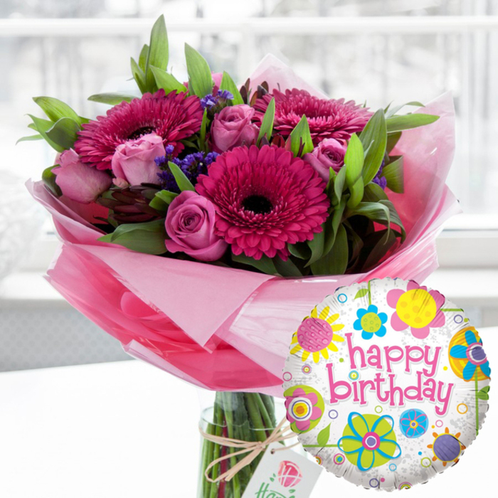 This image showcases a joyful birthday bouquet and balloon pairing, styled with the care of a local florist who knows how people in Epping like to celebrate their special moments. At the centre is a substantial arrangement of fresh flowers presented in a tall, clear glass vase, the stems aligned neatly in the water and subtly magnified by the glass. The bouquet is rich in shades of pink and magenta, anchored by several striking gerbera daisies whose wide, fanned petals glow in vibrant fuchsia tones. Their deep, almost black central discs provide a strong contrast, adding drama and focus. Threaded among them are elegant pink roses, some still tightly furled, others beginning to open into layered rosettes that suggest soft fragrance and timeless romance. Tiny clusters of deep purple filler flowers tuck into the gaps, giving a textured, almost meadow-like feel. Abundant green foliage creates a lush frame, with leaves of varying shapes and textures arching outwards to balance the bright blooms. Around the base of the flowers, translucent pale pink cellophane wraps the stems like a soft cloud, catching reflections from the daylight that filters through, as if from a window looking out toward Epping High Street or Stonards Hill. The vase is finished with a loop of rustic twine, tied into a simple bow that secures a small, off-white gift tag carrying a subtle rose illustration, echoing the craftsmanship associated with Flowers Epping on beville.live. In the foreground, slightly to the right, a glossy Mylar balloon rises, its round shape edging into the frame. The balloon features a white, gently speckled background adorned with cheerful floral and circular motifs in bright yellow, sky blue, lime green, vivid pink and deep purple. Across its centre, the words happy birthday sweep in a bold pink script, making the overall scene feel like a ready-made birthday surprise, ideal for same-day delivery to homes across Epping and the surrounding lanes.