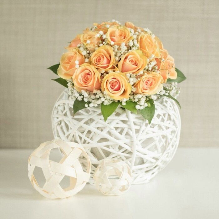 An artfully composed floral arrangement stands on a pristine white table, dominated by a rounded bouquet of around two dozen peach and apricot roses, each bloom placed so closely together that they form a generous, cushion-like dome. The roses vary slightly in openness, some still tightly curled while others gently unfurl to reveal warm, golden-orange centers that gradually soften to pale, creamy outer petals, giving the bouquet a rich, sunlit warmth. Threaded between these roses are airy clusters of white baby's breath, their countless tiny flowers adding feathery volume and a sense of lightness that contrasts beautifully with the denser rose heads. Beneath the blossoms, hints of dark green foliage peep through, offering a natural frame and drawing the eye into the heart of the design. The bouquet is supported by a distinctive spherical vase created from slender white strands woven into an open lattice, rather like an oversized, perfectly shaped string ball, its delicate crisscross pattern lending a contemporary, handcrafted feel. Resting on the smooth tabletop in front of the main vessel are two smaller decorative spheres made from the same white material; one is larger, with panels reminiscent of a minimal, monochrome football, while the other is smaller and slightly irregular, adding a touch of playfulness and visual rhythm. Behind the arrangement, a soft light beige wall with gentle vertical texture provides a calm, neutral backdrop that allows the warm peach tones and detailed weaving to stand out. The lighting is diffuse and natural, suggesting daylight streaming into a tidy Epping living room or boutique along the High Street, where such a centerpiece might grace a coffee table for a birthday gathering or an intimate engagement celebration. The scene communicates both serenity and quiet joy, like something a dedicated local florist at Flowers Epping would create for an elegant home styling or special event in and around the town.