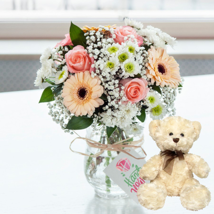 On a sleek white tabletop, bathed in bright yet gentle natural light, a romantic anniversary gift combination is carefully staged, as if ready to be delivered to a home near Epping Station or a quiet cottage on the edge of Epping Forest. The focus of the scene is a generously rounded bouquet arranged in a clear glass jar-style vase, its straight stems visible through the water and neatly aligned by a skilled florist. Soft pink roses, some in tight buds and others more open, mingle harmoniously with peach gerbera daisies that feature deep, chocolate-brown centres. These warm tones are brightened by cushiony clusters of white and lime-tinted chrysanthemums, whose small petals catch the light, and by delicate sprays of baby's breath that float between the larger blooms like tiny white stars. Wrapped around the neck of the vase is a length of natural twine tied in a simple bow, introducing a rustic, homely touch. Suspended from the twine is a petite white tag, decorated with a stylised pink heart or jewel symbol and flowing green script that suggests the words are handwritten, lending a sense of intimacy and care. Just to the right of the flowers, a plush cream teddy bear sits upright, its fur appearing thick and fluffy, inviting to the touch. The bear's dark bead eyes, soft brown nose, and finely tied brown satin ribbon form a friendly, reassuring expression, reminiscent of childhood comfort. The background is softly out of focus, a wash of pale tones hinting at a sunlit interior in Epping, ensuring all attention remains on this charming pairing. Together, the bouquet and bear embody tenderness and appreciation, perfectly suited to anniversaries, romantic gestures, or heartfelt thank-you gifts delivered by local florists.