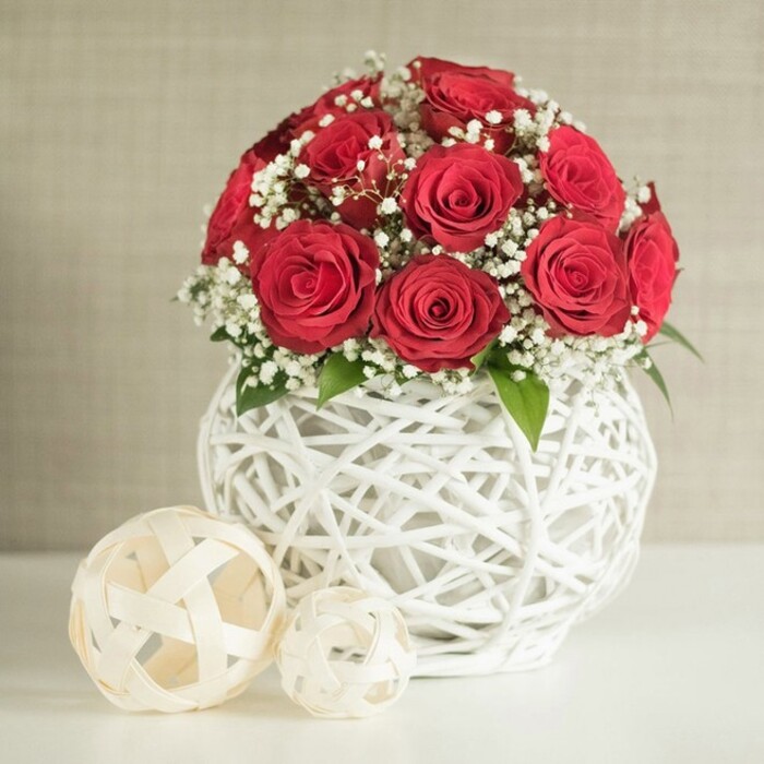 At the centre of this serene interior scene is a lush, spherical bouquet of deep red roses, carefully composed to create a smooth, rounded silhouette that feels perfect for a wedding table or romantic dinner in Epping. Around a dozen to fifteen roses, all in full, velvety bloom, press closely together so that their spiralled petals form a rich, almost quilted surface of crimson. Between these passionate blooms, clusters of tiny white baby's breath emerge, their many miniature flowers looking like soft clouds tucked into the arrangement, lightening the overall effect and providing a gentle contrast. Small glimpses of dark green leaves can be seen beneath the roses, grounding the bouquet in natural freshness and recalling the foliage-lined paths around Epping Forest. The flowers are gathered into a unique round vase, pure white and distinctly woven, as though made from delicately intertwined rattan or wicker. The open lattice design allows light to slip through, casting subtle shadows and giving the vase an airy, handcrafted feel, similar to decorative pieces you might find in local Epping boutiques. The arrangement rests on a smooth white tabletop that reflects a hint of light back onto the vase, maintaining a crisp and uncluttered setting. To the left of the main piece, two decorative woven balls in a soft cream or natural tone echo the lattice pattern of the vase, the larger sphere placed slightly nearer, with a smaller one nestled behind it, adding depth and visual rhythm. The backdrop is a gentle, neutral wall in light beige, its slightly textured surface softly out of focus, helping the vivid red roses stand out. Overall, the image conveys an atmosphere of quiet sophistication and romance, ideal as inspiration for wedding centrepieces or intimate celebration flowers delivered locally across Epping.