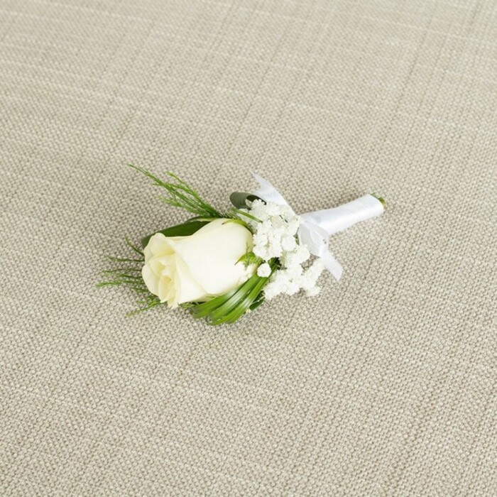 A single white rose boutonniere is artfully arranged at a diagonal angle on a softly textured, light beige fabric, the weave resembling natural linen often seen on rustic-chic wedding tables in Epping venues. At the centre of the composition is a compact, creamy white rosebud, its petals tightly layered yet velvety, with just a hint of ivory along the inner curls. The bloom appears freshly cut, with a subtle sheen that suggests coolness to the touch, and a faint glimpse of green sepals at its base adds a natural transition into the stem. Embracing the rose is a delicate surround of white baby's breath, their tiny clustered blossoms floating like a fine mist, giving the arrangement a gentle, cloud-soft frame. Interspersed among these are slender, deep green leaves and a few feathery, fern-like sprigs, which introduce height and dimension while recalling foliage gathered from leafy corners near Epping Forest. The greenery is carefully positioned to cradle the rose without obscuring its shape, adding structure and a lively garden freshness. All stems are gathered and meticulously wrapped with a narrow strip of pure white satin ribbon, the wrapping smooth and even, running the length of the exposed stems. Just below the floral head, the ribbon is finished in a neat, understated bow, its small loops perfectly balanced, suggesting the measured movements of an experienced wedding florist. Soft, diffused lighting glides across the scene, accentuating the subtle shadows within the rose petals, the airy texture of the baby's breath, and the fine crosshatch pattern of the fabric beneath. The image conveys quiet sophistication and heartfelt romance, an ideal boutonniere for a groom arriving at Epping's local church or registry office, symbolising purity, commitment, and timeless style.