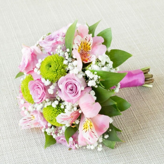 This beautifully hand-tied bridal bouquet, photographed diagonally upon a light ivory linen reminiscent of elegant Epping wedding venues, radiates grace and contemporary charm. At its heart lie enchanting pastel pink roses-some just beginning to unfurl, others in full bloom-each rose boasting velvety petals that spiral towards their delicate centers in a celebration of natural beauty. Nestled harmoniously are vibrant lime-green button chrysanthemums, their tidy pom-pom shapes creating a striking contrast in both colour and tactile appeal. Soft pink alstroemeria, featuring slender petals adorned with subtle yellow highlights and intricate maroon speckling, lend an exotic allure unique to the finest local floral creations. Wispy white sprigs of baby's breath (gypsophila) intermingle, lending a gossamer delicacy and softly blurring the bouquet's edges, recalling a gentle morning walk along Epping High Street. Lush, dark green leaves peek throughout as a vibrant backdrop, imbuing the bunch with structure and a sense of freshness as if just gathered from a woodland border near Epping Forest. The entire bouquet is bound together at the stems in a rich, silky magenta ribbon-its sheen a nod to celebratory joy-secured neatly to enhance both form and comfort for the bridesmaid's grip. The gentle lighting flatters each bloom, allowing texture and nuanced hues to shine, making this bouquet an ideal choice for Essex weddings, spring fêtes, or anniversaries, showcasing the creative flair and dedication of Epping's expert florists.
