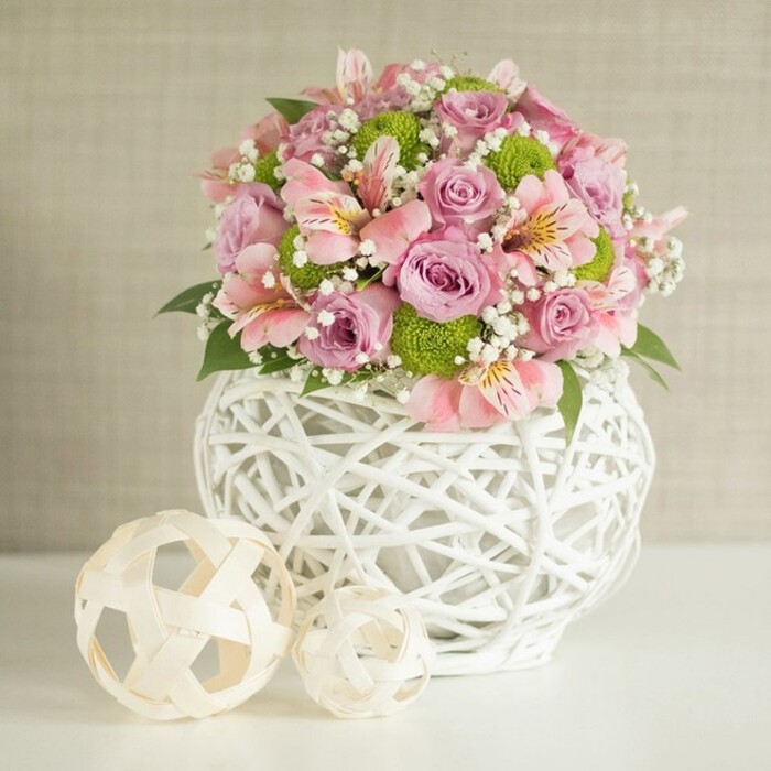 The scene captures a striking floral arrangement, abundant with elegance and freshness, illuminated against a softly textured beige backdrop suggestive of Epping's welcoming interiors. The bouquet is a harmonious medley of full-bloom pastel pink and lavender roses, each unfurling soft, velvety petals that catch the light. Scattered among them are delicate pink alstroemeria lilies, recognisable by their slender, trumpet-shaped blooms and painterly inner striping-details that add subtle complexity. Small, spherical lime green button chrysanthemums nestle in clusters, bringing playful vibrancy and a lively celebration of spring colour. Wispy sprays of white baby's breath float gracefully within the bouquet, lending it a light, ethereal aura. Framing the flowers are deep green leaves, providing contrast and depth, much as Epping Forest frames the town. This lush ensemble rises from a stylish, rounded white vase-an art piece in itself, designed with an open-lattice basket weave reminiscent of crafts found in Epping's artisan shops. The vase provides visual grounding while remaining airy and modern. Two decorative, pale woven spheres rest to the side, their textures and shapes mirroring the vase and inviting thoughts of rustic charm found in local homes. Set upon a clean white table and perfectly lit to highlight every detail, this arrangement is ideal inspiration for weddings held at nearby Epping venues or as a refined home décor touch that echoes the town's unique blend of heritage and innovation.