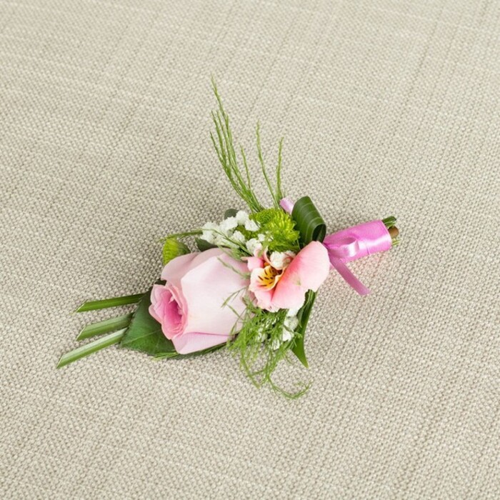 A meticulously composed boutonniere lies diagonally across a light, subtly textured beige fabric-an arrangement radiating the quiet elegance often found in gatherings at local Epping venues. Its focal point is a pristine, softly blushing pink rosebud, tightly furled and glowing with freshness, its centre a deeper pink that fades gently outward. Complementing the rose is a lighter pink or peach-toned blossom-likely Alstroemeria-its petals unfolding to reveal a golden yellow throat delicately marked with fine veins, adding layers of visual and emotional interest. Sprays of fluffy white baby's breath float throughout, evoking a sense of airiness and recalling English garden mornings. Lively pops of vivid green, shaped as miniature cushion mums, nestle within the bouquet, drawing the eye much as Epping's green spaces shape the landscape. The foliage includes a balanced mix of sleek, narrow leaves and broader, darker greens; some are artfully folded at the base, creating depth and support, while others curve upward with a light, feathery touch, reminiscent of ornamental ferns. Wrapping the densely bound stems at the top, a wide, dark green leaf adds both visual interest and stability. The finishing touch is a lustrous fuchsia satin ribbon, meticulously tied in a petite bow, its bright sheen offering a cheerful, sophisticated contrast against the natural hues. The entire design is set against a woven linen reminiscent of traditional Epping homes, its surface picking up gentle highlights in the soft daylight. Perfect for weddings, proms, or cherished celebrations in Epping and the surrounding villages, this florally rich accessory reflects both the skilled hand of its florist and the heartfelt occasions it is chosen for.