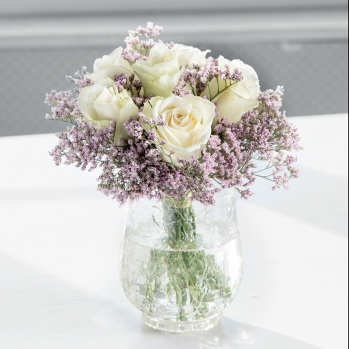 A luxuriously full bouquet of creamy white roses and lavender-tinted statice is captured here in a moment of tranquil beauty, poised in a tall, clear cylindrical vase on a smooth white tabletop. The roses, around a dozen in number, are a soft, almost luminous ivory, their petals layered in generous spirals that appear both plush and delicately thin. Each bloom is at that ideal stage of openness-no longer tight buds, yet not fully blown-so they form a rounded, balanced dome that speaks of classic florist craftsmanship. Between and around these central flowers, wisps of pale lilac statice weave in and out, their many tiny blossoms clustering like a filigree mist. The contrast between the velvety rose petals and the papery, textured statice adds depth and subtle drama without disturbing the arrangement's calm character. Through the transparent glass, you can see the cleanly cut green stems, gathered neatly and immersed in clear water that gleams as it catches the ambient light. The scene is bathed in soft natural daylight from an unseen window, creating gentle highlights along each petal edge and faint reflections on the glossy white surface below. The background remains deliberately blurred, suggesting a bright, quiet interior-perhaps a sitting room in an Epping home overlooking the greenery near Swaines Green or a peaceful corner in a local café. Crafted with the sensibility of an experienced florist at Flowers Epping, the bouquet feels perfectly suited for occasions that ask for refinement without excess: an elegant anniversary surprise, a condolence gift offering calm and comfort, or a sophisticated centrepiece for a small family gathering in the Epping area.