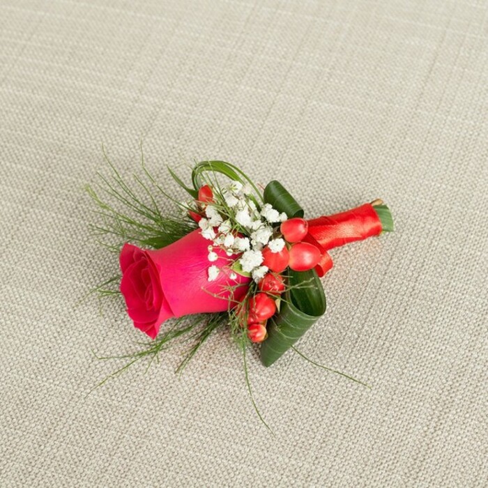 Displayed on a neutral, light beige fabric with a gently textured, woven surface, this elegant boutonniere draws the eye with its balance of rich colour and fine detail, much like the thoughtful wedding flowers chosen by couples across Epping. At the centre is a vibrant fuchsia rose, its petals compact and spiralled at the core, gradually unfolding into a structured, velvety bloom that suggests freshness and subtle perfume. Encircling this focal flower, delicate clusters of white baby's breath create a soft, airy border, each tiny blossom offering a point of light against the deeper hues. Tucked among these white sprays, round red hypericum berries shine with a glossy surface, adding warmth and a festive, almost jewel-like accent. Slender, wispy fronds of fern-like greenery extend outward, giving the piece an organic sense of movement, while broader, darker green leaves are artistically curved and folded to provide a supportive collar that enhances the rose's shape. The stems are pulled together and meticulously wrapped in a smooth red satin ribbon, which is tied firmly near the base to form a neat, understated knot. The ribbon's sheen plays gently with the lighting, echoing the blush of the rose and the glow of the berries. Captured from a slightly overhead angle, every layer of texture-from the linen-like background to the structured foliage-is clearly defined. This small floral accessory feels perfectly suited to a groom, groomsman, or prom guest walking through Epping town centre, a refined touch of romance and ceremony that complements a tailored jacket or formal suit.
