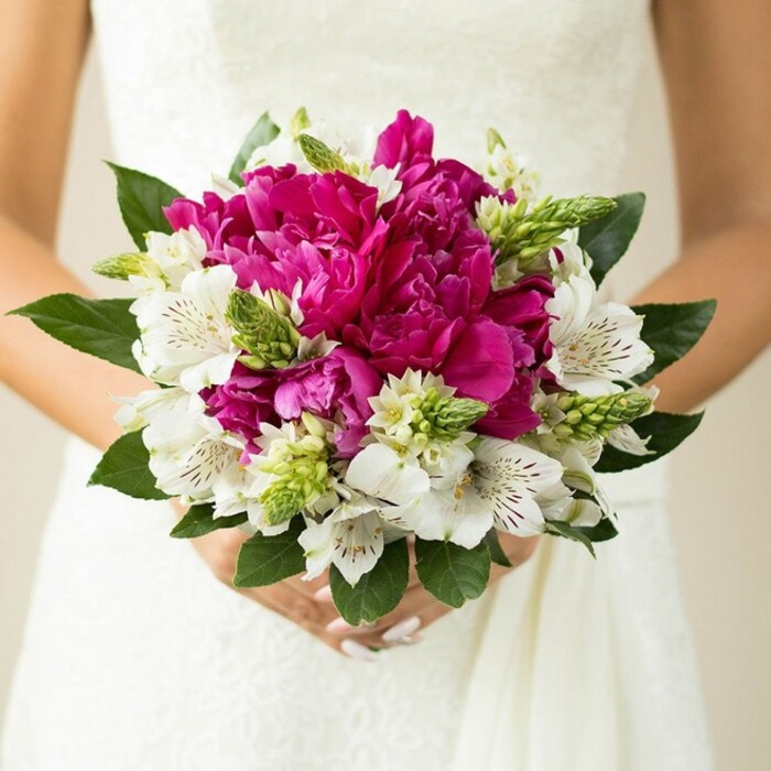 In this close, intimate shot, a bride holds a compact, rounded bouquet just in front of her midsection, the white lace and smooth fabric of her gown providing a gentle, neutral backdrop. At the centre of the arrangement sit large, fully open peonies in a striking fuchsia and magenta palette, their many ruffled petals curling and overlapping to form a lush, velvety core that immediately draws the eye. Nestled among these dramatic blooms are clusters of white alstroemeria, each delicate flower displaying subtle maroon or wine-coloured flecks within its throat, like tiny brushstrokes that add refinement and depth. Threaded through the composition are upright, spiky green buds and textural sprigs, probably Ornithogalum or similar, whose fresh lime-green tones brighten the bouquet and whose pointed silhouettes contrast with the softness of the peonies. Encircling the flowers, broad, dark green leaves fan outward, creating a natural frame that grounds the vivid colours and gives the bouquet a hand-gathered feel, as if the stems could have been picked from a well-tended garden near Bell Common in Epping on a sunny morning. The bride's hands are gently clasped around the stems, her nails polished a pale blush pink that harmonises with the romantic palette. The lighting is soft and diffused, eliminating harsh shadows and allowing every texture-from the silk-like petals to the glossy leaves-to be clearly seen. The overall mood is one of elegant celebration: a modern Epping bridal bouquet designed to stand out in close-up wedding photographs, ideal for couples seeking fresh, vibrant florals that still echo timeless romance and seasonal charm.