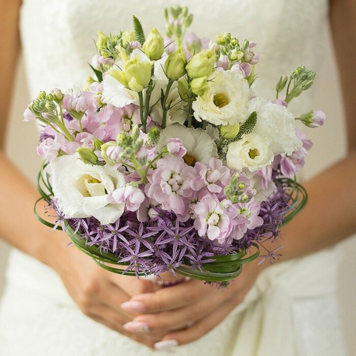 Soft morning light bathes this beautifully composed wedding bouquet, which rests delicately in the gently cupped hands of a bride. The lower edge of the frame offers a glimpse of neatly manicured nails-painted in nude or pale pink-contrasting with the subtle ivory lace or textured fabric of a bridal gown that remains just out of focus in the background. At the core, lush white Lisianthus blooms with ruffled petals and subtle greenish-yellow throats extend outwards, creating an air of refined classicism often sought in Epping's elegant wedding settings. Scattered among these are plump buds and unopened blossoms, their pale greens adding freshness and life. Intertwined through the arrangement, pastel pink and lilac stock flowers fill out the bouquet with a plush, ethereal quality, reminiscent of the wildflowers that bloom each spring along the trails connecting Epping to its charming market town center. A standout feature near the base is a collar of rich purple, spiky Allium flowers, their geometric shapes adding a modern twist to the otherwise romantic design. Above this, glossy loops of bear grass thread through the flowers, providing subtle structure and a touch of whimsy reminiscent of Epping's village greens on a breezy afternoon. The bouquet, tailored to evoke themes of love, freshness, and new beginnings, feels both rooted in tradition and enlivened by contemporary touches, making it a perfect choice for weddings across Epping, whether held in rustic barns or stately manor houses.