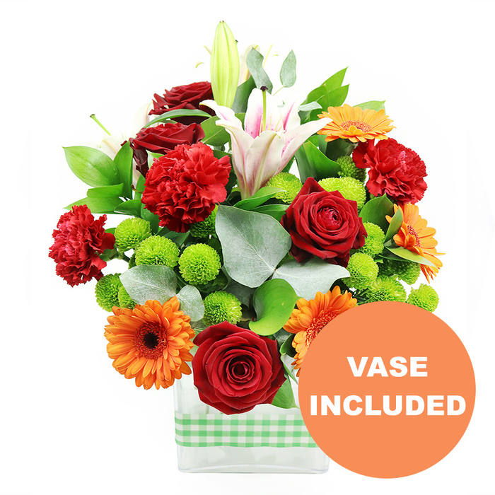 A lush, densely packed floral arrangement is shown in full detail, the kind of vibrant centrepiece that could brighten a breakfast table in an Epping cottage or a modern apartment overlooking the High Road. Deep red roses, fully formed with layered, velvety petals, nestle alongside ruffled red and orange carnations, their frilly edges catching the light and creating a soft, textured halo around each bloom. Bright orange gerbera daisies with dark, almost black centres fan outwards, their flat petals arranged like spokes, lending a bold, contemporary energy. Between the warmer tones, clusters of lime-green button chrysanthemums appear as small, perfectly rounded spheres, giving a lively, bubbly contrast that keeps the design feeling crisp and modern. Elegant white Oriental lilies stretch taller than the other flowers, some star-shaped and fully open to reveal slender pink stripes running through the petals and prominent stamens at the centre, while others remain in smooth, tapering buds ready to open over the coming days. The bouquet is anchored in a clear square-edged glass vase, its transparency showcasing the green stems and the clean line of water within. Around the base, a light green and white gingham ribbon wraps snugly, contributing a cheerful, country-style accent that feels right at home in the leafy surroundings of Epping and its nearby villages. Rich, glossy foliage in deep shades of green, along with soft, silvery eucalyptus leaves, frames the flowers and fills the gaps, adding depth and a gentle, natural movement to the arrangement. In the lower right corner, partly overlapping the vase, an orange circular graphic clearly displays the words VASE INCLUDED in bold white capitals, making it evident that this is a complete, ready-to-display floral gift, ideal for celebrations, thank-yous, or welcoming guests in Epping.