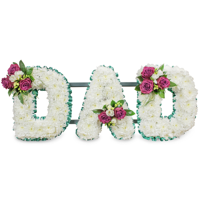 Presented on a plain white backdrop, this memorial piece forms the word DAD in large, three-dimensional capitals, each letter densely packed with pristine white chrysanthemums that create a full, cushion-like face. The compact chrysanthemums form a cloud of soft petals, giving the letters a gentle volume and an impression of calm repose. Each letter is outlined with a pleated emerald-green ribbon that defines the shapes with a formal, elegant border and a subtle textural contrast to the smooth white blooms. A muted dark-green or grey horizontal support bar runs behind the letters, suggesting the robust mount used for display at a funeral or graveside service. Small, carefully placed accent clusters bring colour and personalisation: the first D bears a grouping of deep magenta roses and small white freesia on its upper-left curve; the A holds its cluster tucked into the lower central V; the final D echoes the first with a similar spray on the upper-right curve. The magenta roses introduce a warm, heartfelt hue while the freesia adds delicate fragrance and bright white highlights against the darker foliage. As an arrangement crafted for solemn remembrance, it reads as an intimate tribute suitable for services around Epping - from chapels on the High Street to graveside ceremonies near Bell Common or Theydon Bois. Prepared with the care of a local workshop, this DAD lettering balances traditional respect and personal warmth, offering a quiet but unmistakable message of love and farewell - often chosen by families seeking a dignified, personalised expression of sympathy from Flowers Epping.