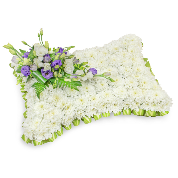 A carefully formed rectangular cushion of flowers sits against a stark white field, its soft three-dimensional presence enhanced by subtle shadowing beneath. The foundation is an even, dense covering of small white chrysanthemums whose rounded heads create a plush, cloud-like texture that suggests peace and purity; their compact arrangement produces a serene, unbroken expanse of white. Encircling this expanse is a thoughtfully pleated, light lime-green ribbon trim that follows the pillow's contours, the neat folds offering a gentle contrast and a touch of renewal. Diagonally placed at the upper-left corner, a composed spray provides the arrangement's focal point: crisp white lisianthus intermingled with deep purple lisianthus blooms and tight buds, the purple tones introducing dignity and quiet sorrow against the white backdrop. Interspersed fern fronds and broad, dark-green leaves bring depth and natural rhythm, while a darker green wrap at the base neatly holds the stems together, revealing the careful hand of the florist. The visual impression is calm and consoling-an arrangement that reads like a soft embrace and is appropriate for funeral settings, memorial services, or placement at a graveside near the High Street or by the town's station. Soft lighting emphasizes the texture of petals and ribbon and suggests an atmosphere of respectful remembrance, conveying solace when words alone cannot comfort those who gather to honour a life.