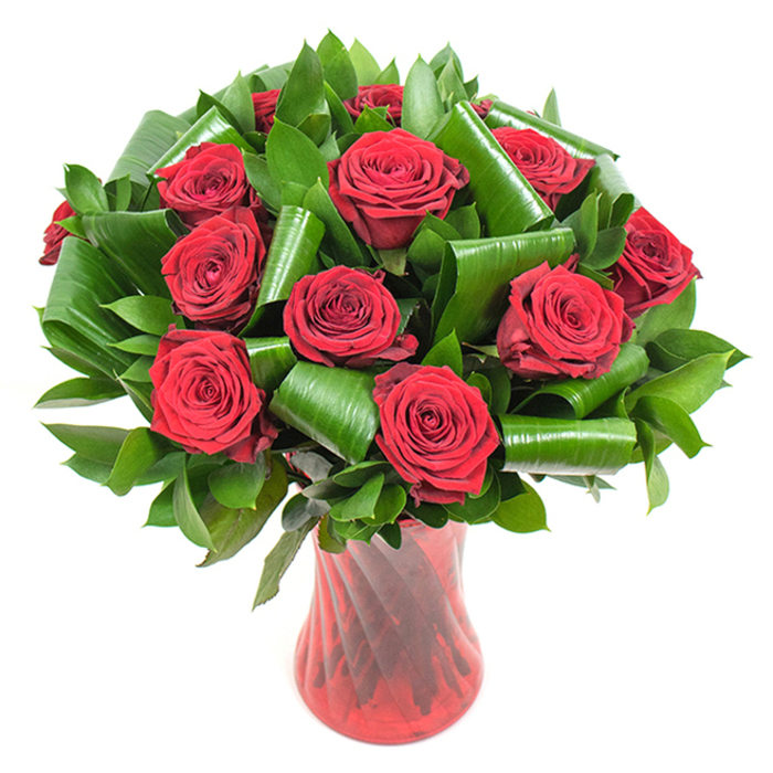 In this image a sumptuous bouquet of roughly a dozen deep crimson roses is composed with an eye for classical romance and contemporary structure: each rose opens fully, revealing tightly wound, velvet-thick petals whose colour graduates from saturated crimson to a near-burgundy depth in the centre. The blossoms are framed by an abundance of dark green foliage-slick, lance-shaped leaves providing a compact, verdant backdrop, while larger, lustrous leaves have been creatively rolled and folded into smooth spirals and arcs that cradle individual blooms. Those rolled leaves give the arrangement an architectural silhouette, looping like green calligraphy around the rose heads and adding volume without obscuring the flowers. The stems settle into a tall, slender ruby-red glass vase that is translucent with a faint ribbed or twisted texture; its vertical pattern refracts light and complements the roses' rich tones while its narrow neck gives the display an elegant, upright carriage. Set against a pristine white background with gentle, even illumination, details such as petal veining, subtle sheen on the greeneries, and the vase's glossy finish are all crisply visible. Imagine receiving this as a gift delivered from a local shop near Epping tube station or placed on a mantel after an evening walk through Epping Forest-the arrangement reads as an icon of romantic intent, suitable for anniversaries, Valentine's Day, or any occasion when enduring passion and refined design are desired.