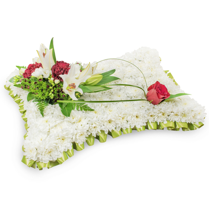 Presented against a neutral white ground, this rectangular floral pillow is a carefully constructed expression of sympathy, its whole surface composed of thousands of tiny white chrysanthemum heads packed tightly together to read as a single, soft expanse. The texture is deliberate-plush, uniform, and quietly luminous-so that the eye is drawn immediately to the subtle pale lime-green satin border that has been neatly pleated and applied around the edge, suggesting the gentle pleats of fabric. On the upper left a composed cluster of accent blooms lifts from the cushion: two fully open white Oriental lilies with their pale yellow stamens exposed, an unopened lily bud offering a vertical line, and clusters of deep burgundy spray carnations whose tight, ruffled petals provide a pleasing counterpoint in tone and shape. A mixture of feathery fern fronds and broader glossy greens frames this cluster and adds natural depth. From that grouping two slender, arching lengths of green bearing grass or salal trace a graceful curve across the cushion, culminating in a single deep red rosebud resting on the upper right-the rose's slowly unfurling petals a private symbol of affection and remembrance. The colour story of crisp white, sombre red and fresh green is restrained and respectful; this is the kind of floral tribute families around Epping Forest and the High Street might choose to express condolences at a wake or funeral service, offered with the quiet attention to detail of an experienced local florist.