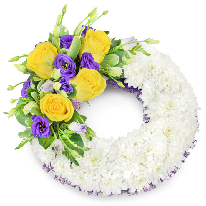 A carefully crafted circular funeral wreath, photographed against a simple white background, that balances reverence with a gentle touch of colour familiar to families across Epping. The majority of the ring is built from compact, snow-white spray chrysanthemums arranged so closely their overlapping petals form a plush, continuous band - a soft, even texture that reads as peaceful and pure. From the upper-left arc springs a focused spray of bright, golden-yellow roses, their fully unfurled petals radiating warmth, counterpointed by clumps of deep purple lisianthus whose ruffled, cup-like flowers add a velvety, noble contrast. A handful of pale lavender lisianthus buds and fresh green growth nestle among the blooms, giving the composition depth and the quiet suggestion of new beginnings. Complementary foliage - glossy, rounded leaves, some with light variegation, and delicate fern fronds - frames the blooms and lends a natural, garden-fresh backbone to the arrangement. A delicate trim of dark-purple mesh ribbon is visible beneath the chrysanthemum base along both inner and outer edges, a subtle flourish that echoes the purple tones in the main spray and provides a refined finish. The wreath's open centre and symmetrical form underscore its traditional role as a sympathy tribute. This piece, suitable for services at local venues such as St John's or memorials near Epping Forest, is the sort of dignified arrangement we create at Flowers Epping - intended to convey heartfelt condolences and a calm sense of remembrance.