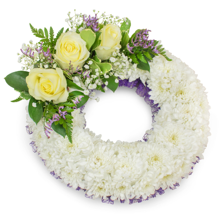 Presented against a stark white backdrop, this circular sympathy wreath has been carefully composed from hundreds of small, immaculate white chrysanthemums to form a dense, uniform ring that feels plush and reassuring to the eye. The tiny chrysanthemum petals layer into a soft, tactile surface that suggests peace and solemnity; the wreath's open centre remains clear, symbolising continuity and unbroken memory. Along the upper-left quadrant a considered cluster of three creamy yellow roses provides a tender focal point-the roses' velvety petals curl outward in quiet bloom-encircled by clouds of white gypsophila and small spikes of pale purple statice or limonium, which lend a restrained hint of colour and a fine, textural contrast. Mixed foliage of glossy green leaves and delicate ferny fronds frames the accent composition, injecting freshness and natural movement. A fine, deep purple ribbon or mesh subtly peeks through the white flowers at both inner and outer edges, adding depth and a refined border to the wreath's silhouette. The lighting is soft and neutral, casting a faint shadow that anchors the wreath on the white surface and reveals the careful craftsmanship of each pinned bloom. Crafted locally by Flowers Epping, this arrangement reflects the kind of thoughtful mourning tribute often selected for services in the town-whether at a chapel on the High Street, a village church near Bell Common, or a graveside in the wider Epping Forest area-conveying respect, comfort and enduring remembrance.