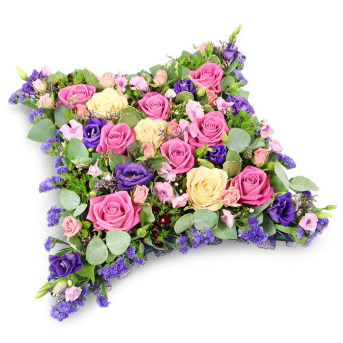 Set against a crisp white backdrop, this elaborate floral cushion tribute appears almost like a soft bed of petals, its pillow-shaped outline neatly rounded at the corners to suggest comfort and rest. The entire surface is tightly packed with roses in varying shades, from striking fuchsia and warm mid-pink to delicate cream and pale yellow, each flower at a slightly different stage of opening to create a layered, three-dimensional effect. Their velvety petals catch the light in subtle highlights and shadows, emphasising the craftsmanship of the florist who arranged them. Bold accents of violet and royal-purple lisianthus are scattered throughout, their gently ruffled blooms lending a rich, almost regal tone that balances the sweetness of the pinks and creams. Around and between the larger flowers, clusters of miniature spray roses and fine, airy lavender-hued filler blossoms, reminiscent of statice or limonium, fill any gaps so that no part of the cushion appears bare. The flowers are cradled by an abundance of greenery: round, soft eucalyptus leaves in muted silver-green tones combine with finer, deeper green, fern-like foliage, creating layers of texture and a cool, refreshing contrast. A subtle shadow beneath the cushion gives the impression it is lightly resting on a tabletop, ready to be carried to a service in Epping, perhaps to St John's or a quiet chapel by the forest. The overall composition feels thoughtful and composed, a floral pillow that communicates love, gratitude, and gentle farewell, much like the bespoke tributes regularly created by experienced local florists for funeral and memorial gatherings across the Epping community.