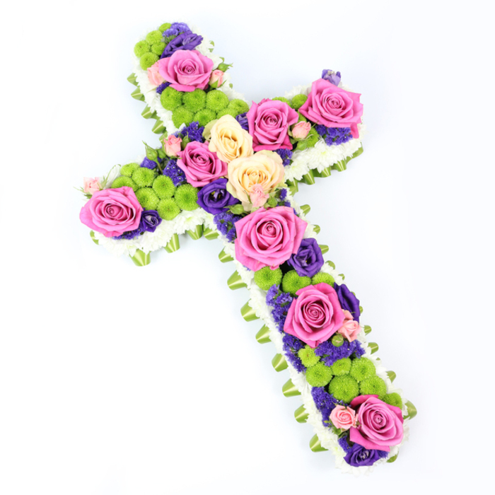 Set against a bright, uncluttered white background, a meticulously crafted floral cross fills the frame, angled slightly from the upper left to the lower right so its three-dimensional form is clearly visible. The craftsmanship is evident in the precise outline formed by a thick border of white spray chrysanthemums, each tiny bloom carefully positioned to create a soft, quilted edge that feels both pure and comforting. From beneath this white border, a subtle band of light green ribbon or foliage emerges, neatly pleated and wrapped around the perimeter of the cross, adding a refined finish and a hint of fresh colour reminiscent of the green verges and parkland around Epping. Within this pale frame, the interior of the cross is alive with texture and colour. Large roses in rich hot pink and softer creamy yellow or peach tones are arranged in gentle clusters, their full, open faces drawing the eye along the length and across the arms. Nestled between the roses are vivid lime-green button chrysanthemums, their round, compact shapes offering a lively contrast, like tiny mossy domes scattered through a garden. Deep purple and violet flowers, probably lisianthus or statice, are threaded throughout, filling gaps and adding shadows of colour that make the arrangement feel layered and deep rather than flat. The lighting is bright and even, allowing you to see every petal edge, every fold of ribbon, and every subtle colour shift. As a funeral or sympathy tribute, this cross feels appropriate for a church or crematorium service in Epping, expressing reverence, remembrance, and a quiet celebration of a loved one's spirit through its harmonious blend of whites, greens, pinks, and purples.