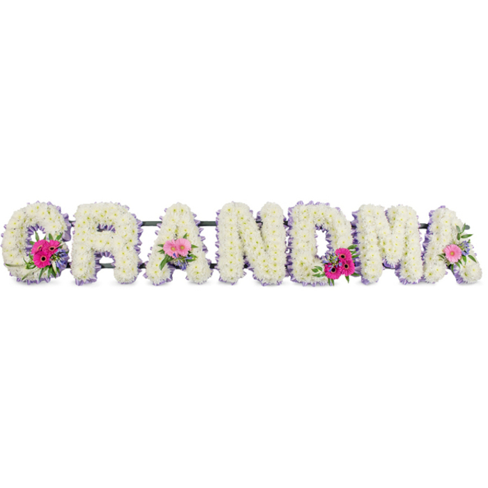 The image shows a beautifully composed funeral flower tribute spelling out GRANDMA, designed as seven bold, upright letters, each one meticulously padded with white chrysanthemums. The blooms are packed closely together, so no gaps in greenery show through, creating an immaculate, snowy surface that feels soft and plush, like a favourite cushion from a grandmother's living room. Around the contour of each letter, a narrow lilac ribbon has been carefully pinned in place, following every curve and corner. This gentle purple outline adds definition, helping the name stand out clearly while also giving a touch of refinement and feminine grace. At three places across the tribute-on the opening G, one of the central As, and the final A-small round bouquets break the field of white. These posies feature cheerful pink gerbera daisies and blushing pink roses, interlaced with sprigs of mixed foliage in varied shades of green, from bright, fresh tips to deeper, woodland tones. The contrast between the pristine chrysanthemums and the playful pink flowers evokes a balance of serenity and cherished, lively memories. Soft, even lighting similar to a calm afternoon over Epping's quiet residential streets illuminates the tribute, casting light shadows that emphasise the three-dimensional form of the letters without appearing harsh. You can picture this tribute resting gently beside other sympathy flowers at a chapel of rest in Epping or being carried with care to a cemetery nearby. The overall mood is one of gentle remembrance, gratitude, and lasting affection, honouring a beloved grandmother with flowers that speak softly when words are hard to find.