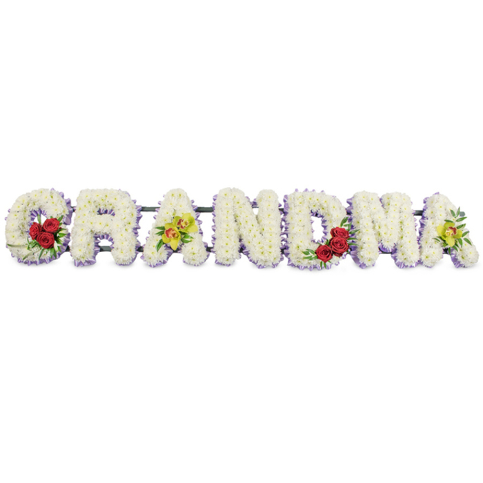 This photograph presents a lovingly made GRANDMA funeral flower arrangement, the word composed in large capital letters that sit in a clear horizontal line, almost floating against a pristine white background. The craftsmanship is meticulous: each letter is entirely covered in small, round-headed white chrysanthemums or similar blooms, positioned so closely together that no gaps in the surface can be seen. The result is a smooth yet softly textured appearance, like a white quilt of petals, gently reflecting the surrounding light. Underneath and just at the edges, a narrow band of pale lavender or soft purple flowers subtly traces each shape, giving a delicate shadow of colour and helping the letters stand out from one another. At the left, the G is embellished along its lower curve with a compact cluster of three deep, velvety red roses, nestled into a bed of glossy green leaves. These roses add emotional depth, suggesting admiration and enduring love. The first A carries two luminous yellow-green cymbidium orchids, their petals open wide, revealing warm reddish-brown markings in the centre that draw the eye and lend a hint of exotic grace. The middle letters R, N and D remain purely white with their lavender underlay, creating a calm, uninterrupted flow across the centre of the tribute. On the M, another trio of red roses is carefully placed in the lower right area, echoing the roses on the G and adding balance, while the final A repeats the pair of chartreuse orchids, uniting the beginning and end with the same distinctive bloom. An unobtrusive dark green or black metal framework links the letters from behind, giving the tribute the strength it would need to be transported to a church or crematorium in Epping or to rest beside the family during a service. The overall arrangement conveys a deep sense of respect and tenderness, a gentle floral statement of gratitude for a grandmother's life and the memories she leaves behind.