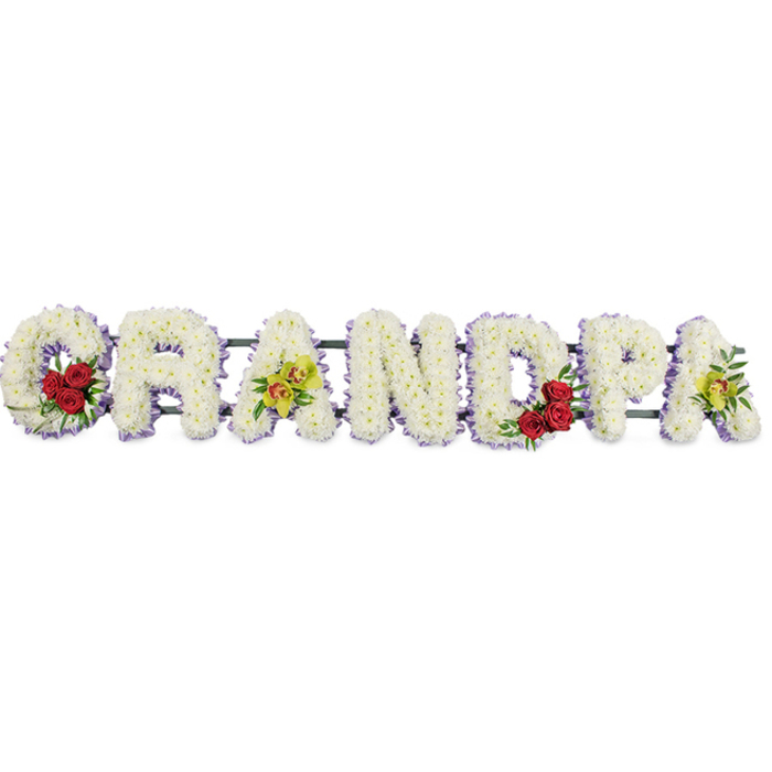 The image shows a carefully crafted funeral tribute that spells out GRANDPA in prominent, horizontal lettering, all set against a smooth, white backdrop that gives the flowers a sense of stillness and clarity. Each letter is formed from tight clusters of small white chrysanthemums, arranged closely together so that no gaps of green or frame show through, creating a dense, pillow-like surface that feels soft and consoling. Around the edges of each letter runs a fine border of lavender or soft purple ribbon, which subtly frames the white blooms and brings a quiet note of colour reminiscent of evening light over the fields around Epping. The florist has introduced pockets of deeper colour that lend the tribute its emotional focus. Nestled in the curve of the capital G is a compact grouping of velvety red roses, interwoven with sprigs of dark, glossy foliage; their rich hue evokes enduring love and heartfelt remembrance. A matching group of red roses is tucked into the rounded section of the D, tying the composition together and giving a gentle sense of balance. On the first A and the last A, small sprays of yellow-green orchids with lightly speckled throats add a note of elegance and individuality, suggesting admiration and the uniqueness of the grandfather remembered. Slim, unobtrusive metal bars run along the back, holding the letters in line so the word can be displayed easily at a funeral service or graveside. This is the sort of bespoke piece a family in Epping might order from a local florist for a final tribute, to sit near the coffin or at the front of the chapel as relatives gather to say goodbye.